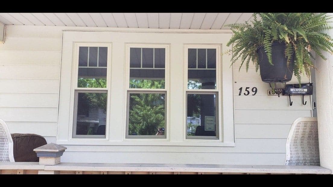 White house exterior with three large windows, address number 159, hanging fern basket, and porch seating including wicker chairs