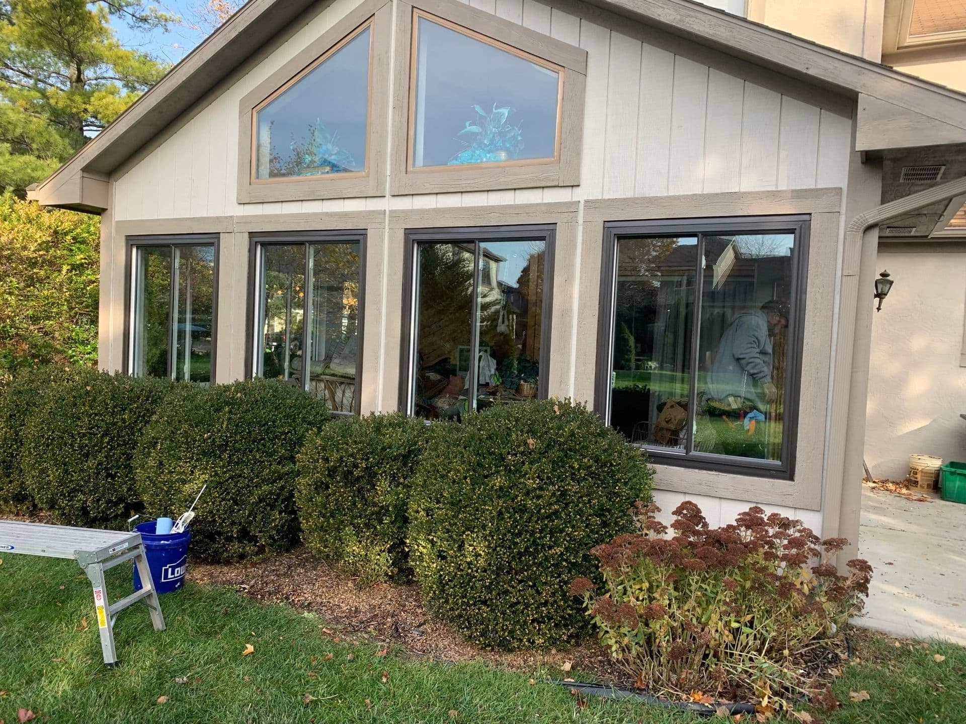 Modern house exterior with multiple large windows and glass doors, manicured hedge landscaping in front, and patio area visible through windows