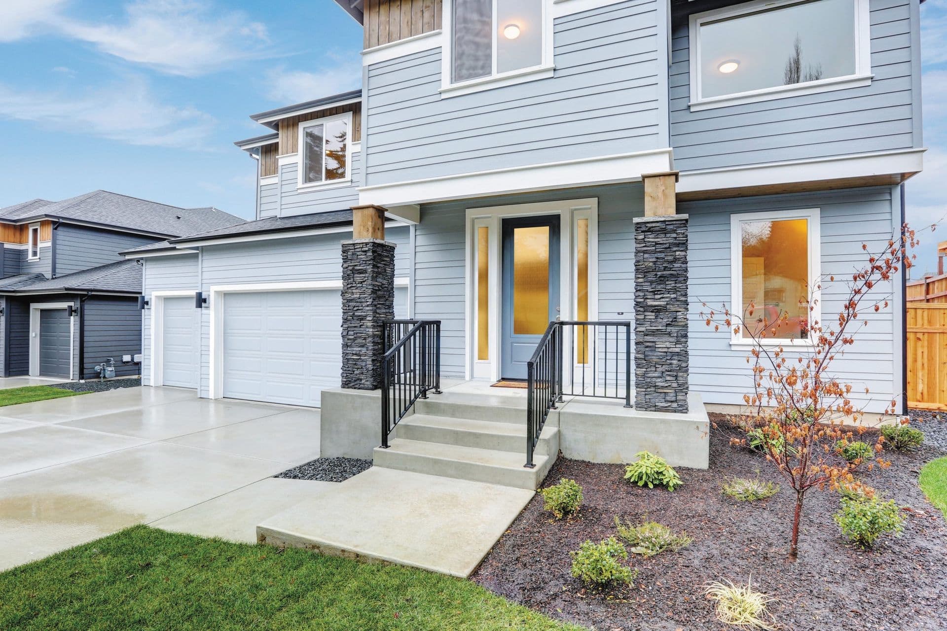Modern two-story home with light blue siding, stone pillar accents, black metal railings, and a two-car garage, with landscaped front yard and concrete driveway