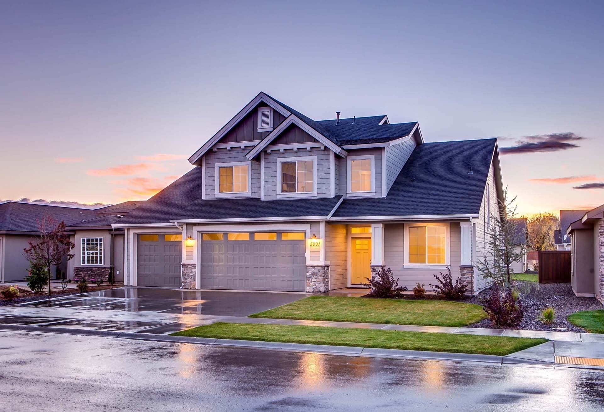 Modern two-story home with white siding and dark roof at dusk, featuring a two-car garage, lit windows, and manicured landscaping