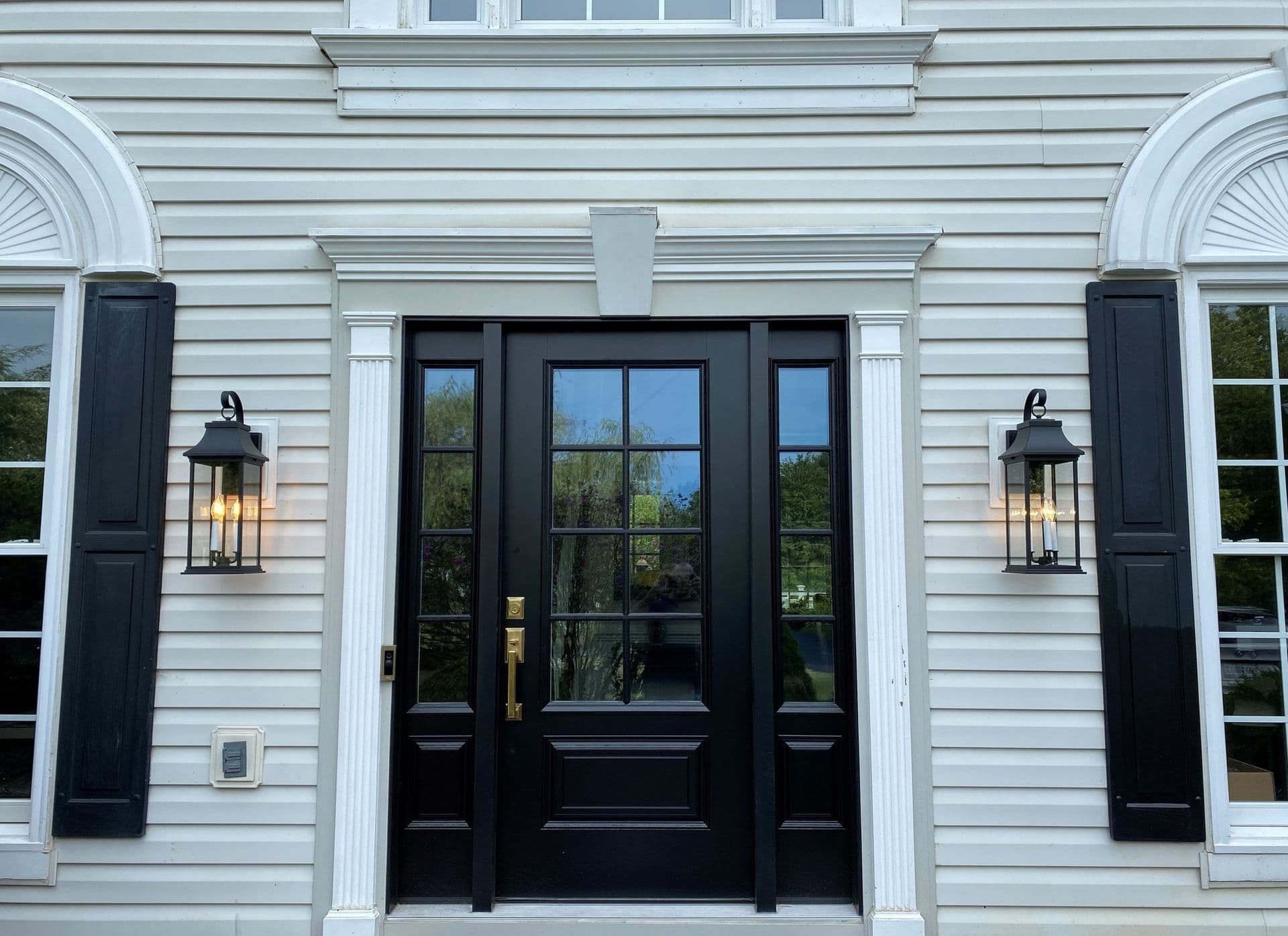 Black front door with sidelights and transom window flanked by white shutters, lantern sconces, and white horizontal siding on residential home entrance